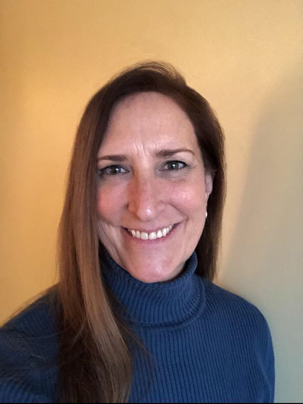 Headshot of Gigi Degerlund Willett smiling, with long reddish brown hair, wearing a blue turtleneck, in front of a yellow wall